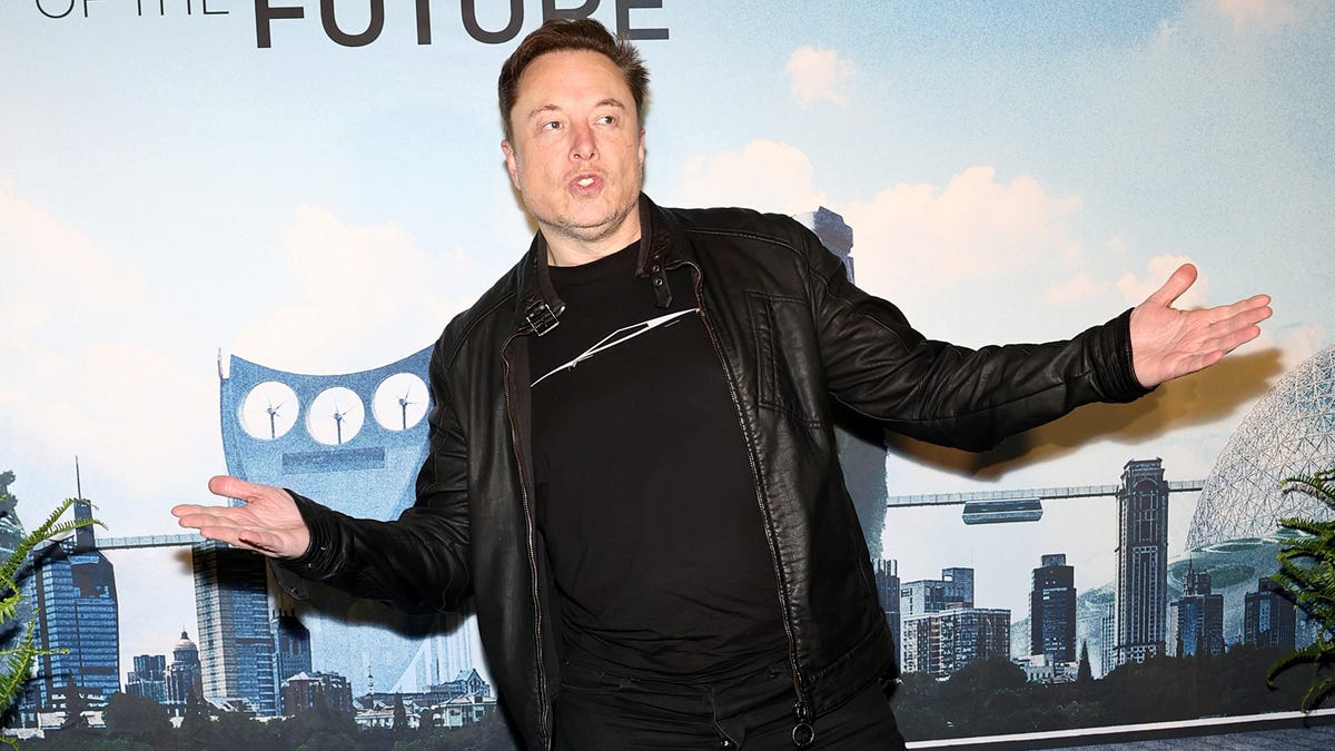 Elon Musk stands in front of a graphical cityscape backdrop with a futuristic robot character, wearing a black leather jacket and gesturing outward with both hands, at the Tesla Robotaxi reveal event.