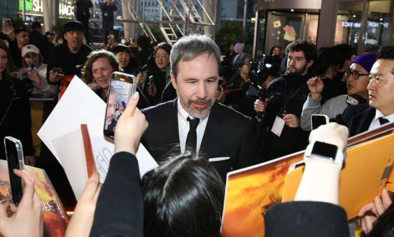 A man in a black suit and tie is surrounded by fans reaching out with posters and a smartphone, taking photos and seeking autographs at a crowded event promoting Villeneuve's next project after Dune