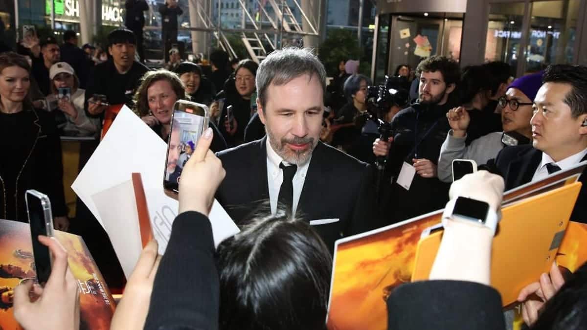 A man in a black suit and tie is surrounded by fans reaching out with posters and a smartphone, taking photos and seeking autographs at a crowded event promoting Villeneuve's next project after Dune