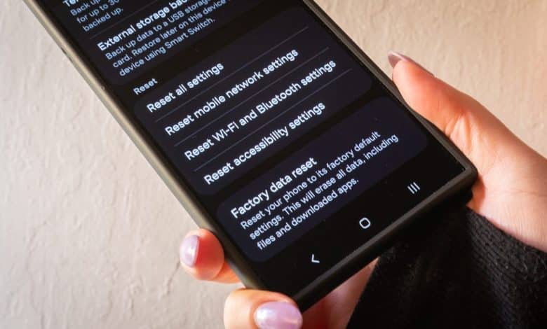 A person holds a smartphone displaying a settings menu with options like "reset wi-fi, mobile & bluetooth," and "factory data reset." The hand has light pink nail polish, and the background is a