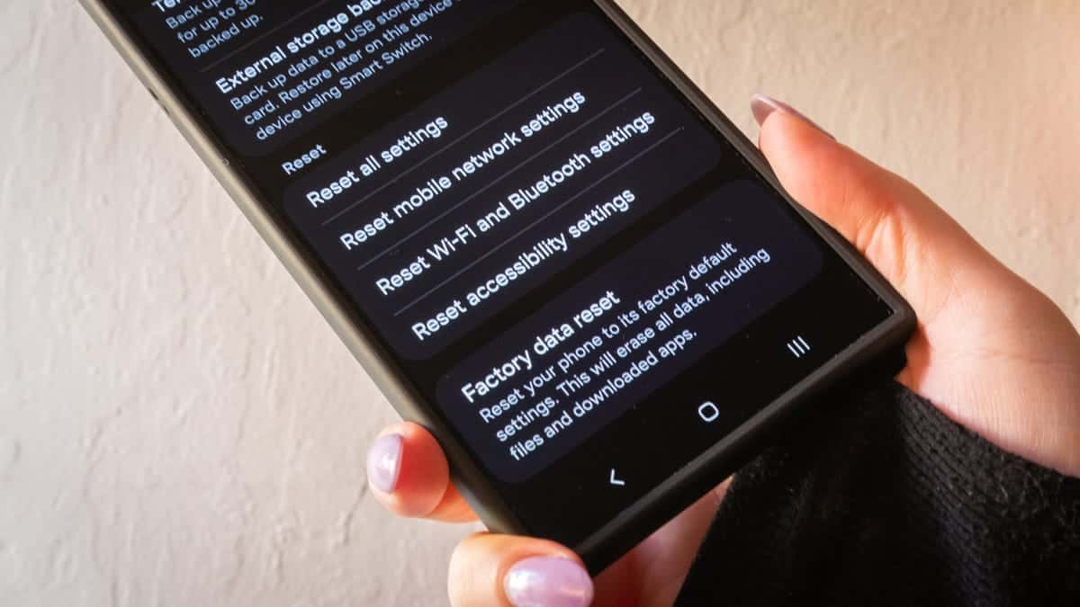 A person holds a smartphone displaying a settings menu with options like "reset wi-fi, mobile & bluetooth," and "factory data reset." The hand has light pink nail polish, and the background is a