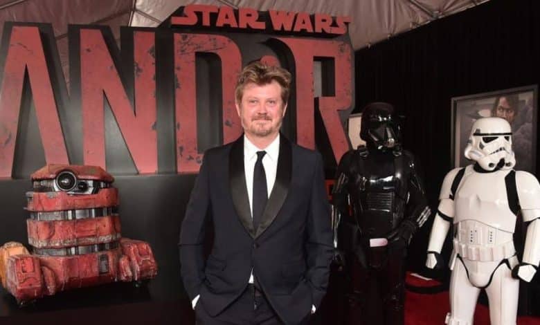 A man in a dark suit stands on the red carpet at the "Andor" premiere, flanked by a stormtrooper and a character in red armor, with the large "Andor