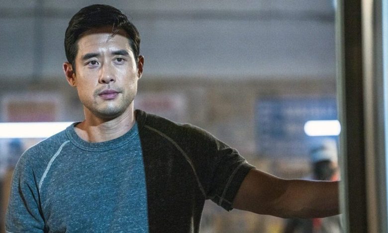 An Asian man in a grey t-shirt stands in a room, holding onto a door frame with a serious expression on his face. He looks focused, with a hint of concern in his eyes as he