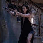 A woman in a black dress, emblematic of John Wick's Ballerina, holds a submachine gun, bracing against a stone wall with intensity in her stance and sharp focus on her face