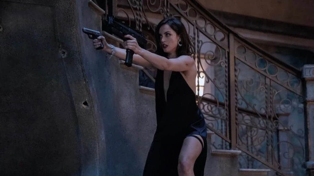 A woman in a black dress, emblematic of John Wick's Ballerina, holds a submachine gun, bracing against a stone wall with intensity in her stance and sharp focus on her face