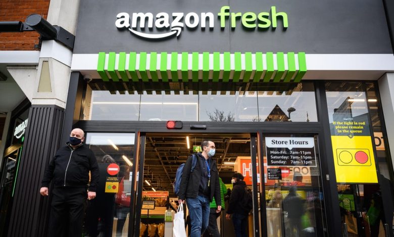 Entrance of an Amazon Fresh store with two men, one exiting and one entering. The facade includes the Amazon Fresh logo and green-striped awning, with posted store hours and a lighted sign showing