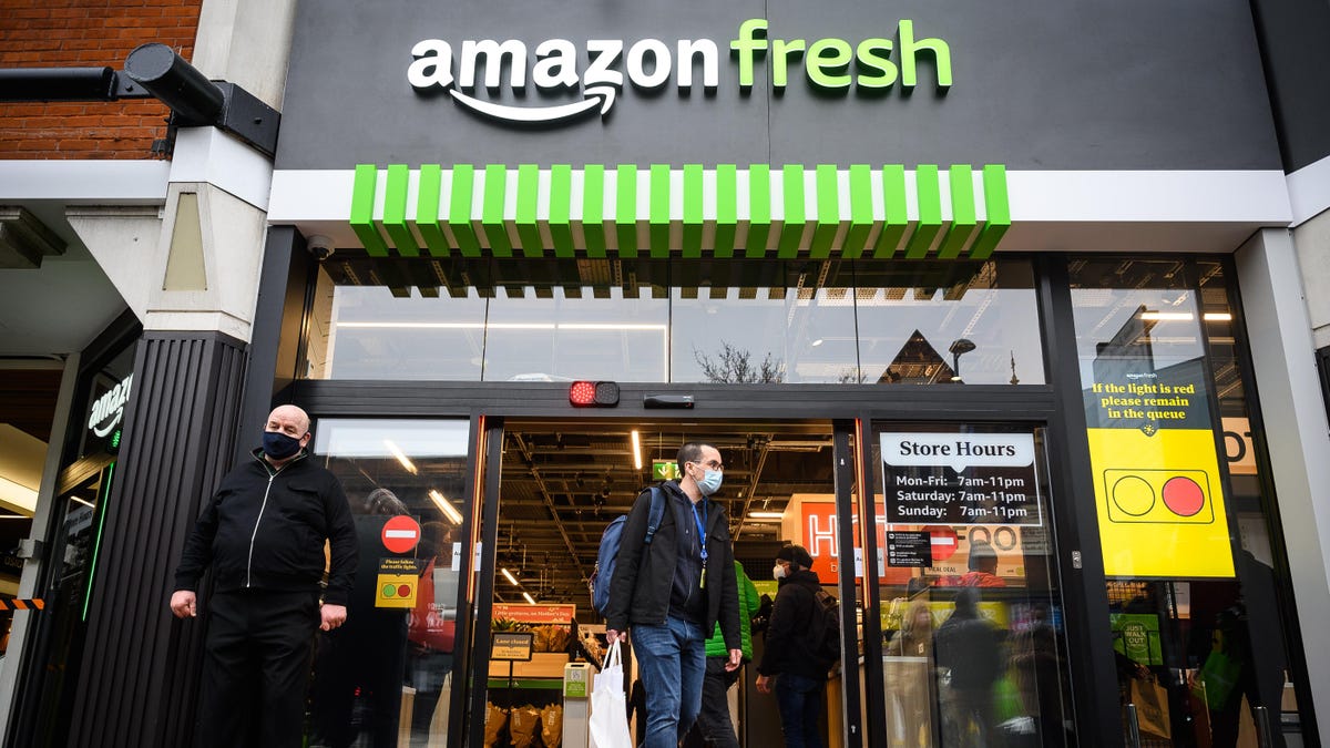 Entrance of an Amazon Fresh store with two men, one exiting and one entering. The facade includes the Amazon Fresh logo and green-striped awning, with posted store hours and a lighted sign showing