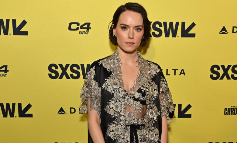 A woman, resembling Daisy Ridley, stands against a branded sxsw backdrop. She wears an elegant, black and silver lace dress, and has short dark hair styled in waves, looking directly at the camera