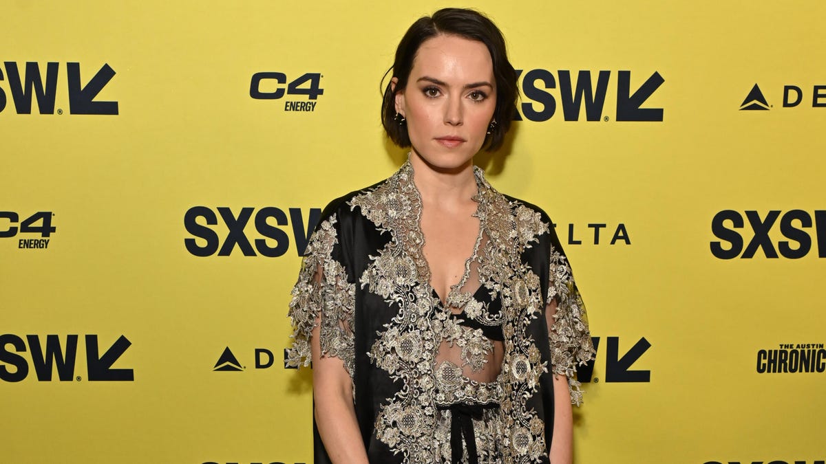 A woman, resembling Daisy Ridley, stands against a branded sxsw backdrop. She wears an elegant, black and silver lace dress, and has short dark hair styled in waves, looking directly at the camera