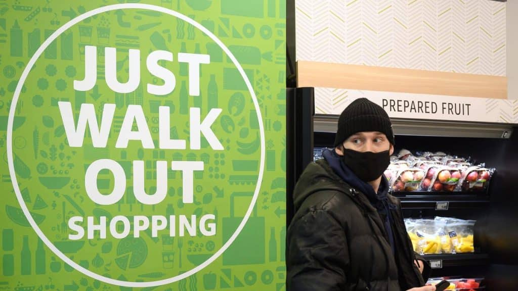 A person wearing a black beanie and face mask stands next to a sign that says "Amazon ends 'Just Walk Out' tech development" in a grocery store beside the prepared fruit section.