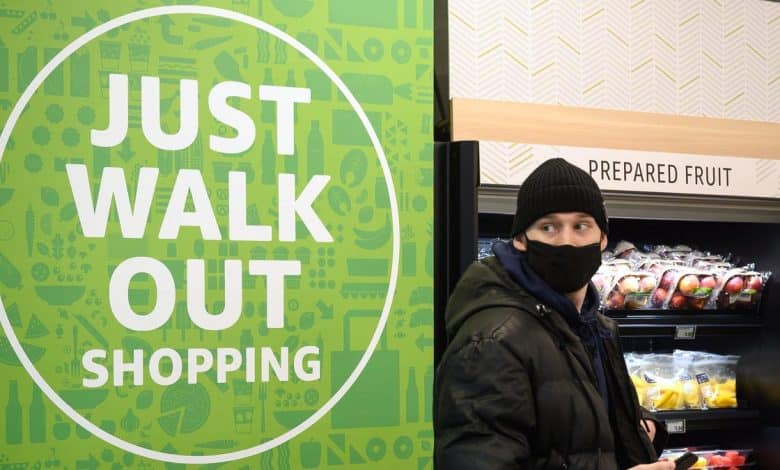 A person wearing a black beanie and face mask stands next to a sign that says "Amazon ends 'Just Walk Out' tech development" in a grocery store beside the prepared fruit section.