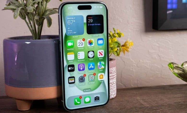 A smartphone in a stand on a wooden surface, displaying a weather app, icons for various other apps including "8 Strategies to Boost iPhone Battery Life", and a cluttered background with a potted plant