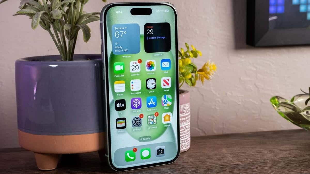A smartphone in a stand on a wooden surface, displaying a weather app, icons for various other apps including "8 Strategies to Boost iPhone Battery Life", and a cluttered background with a potted plant