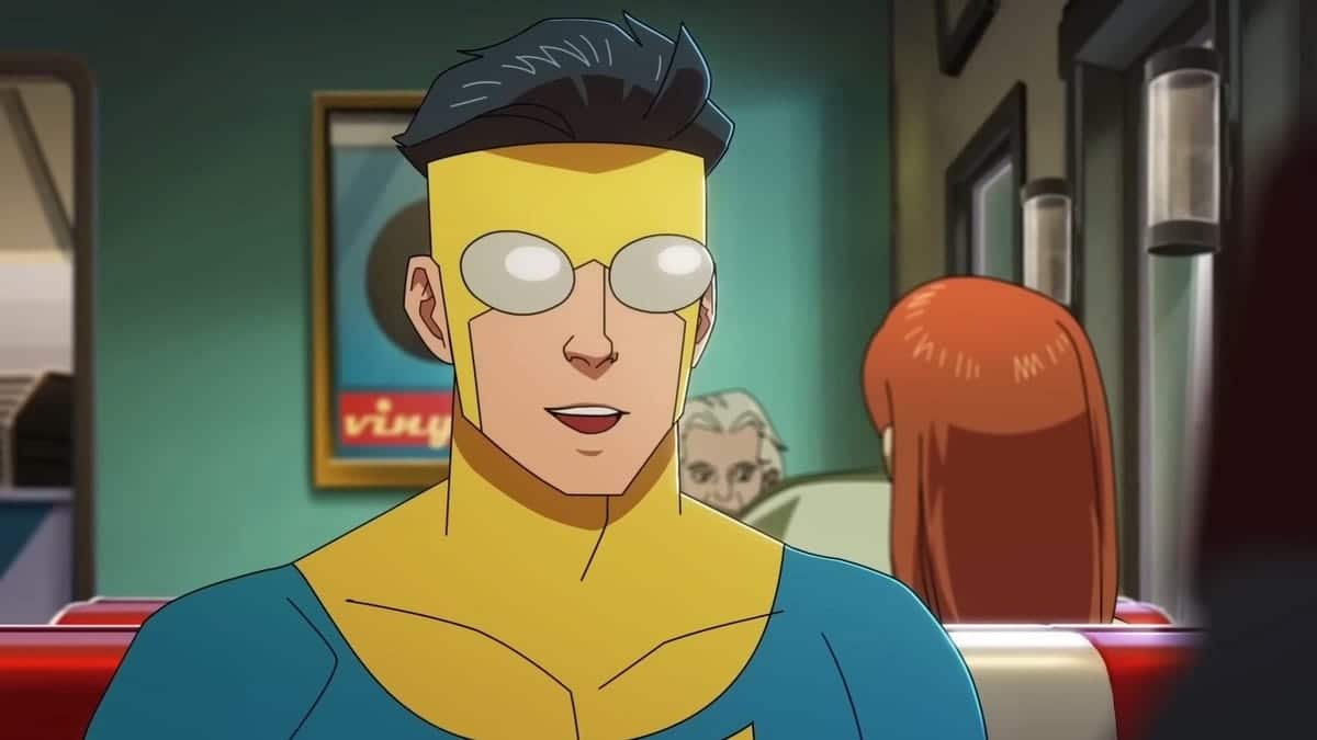 A scene from an animated series showing a young male superhero with yellow goggles, spikey dark hair, wearing a blue and yellow suit, seated, and looking slightly surprised in a room with green