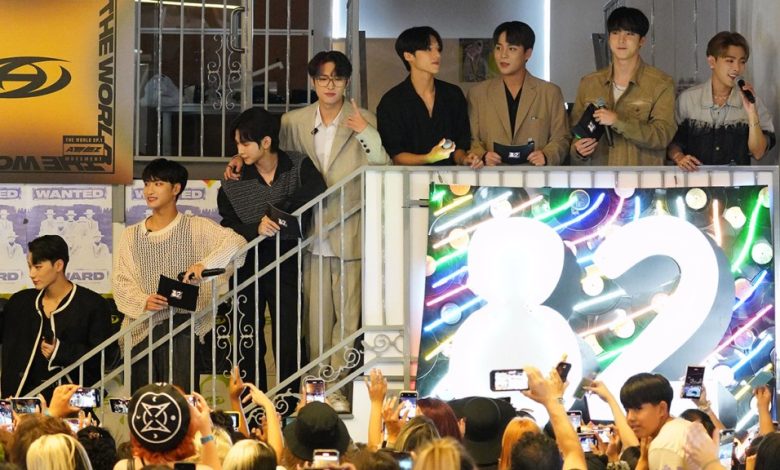 A group of seven young men, likely a boy band associated with Hello82, stands atop a staircase at a public event. They are observed by a crowd below, some holding up cameras. Behind them