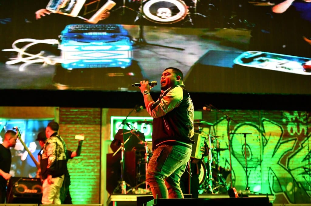 A male singer in a green jacket and torn jeans passionately performs on stage at a concert with his band. Large screens in the background display close-up images of a DJ's equipment, as part of the