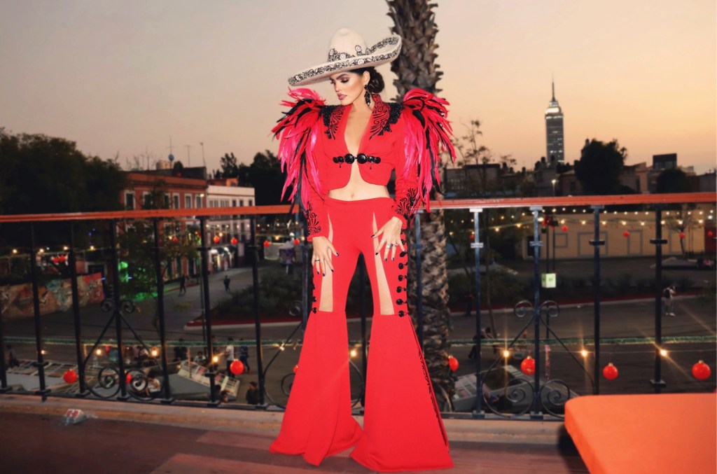 A woman in a vibrant red, flamboyant costume with feather details stands on a balcony at sunset. Her outfit includes a wide-brimmed hat and she's accessorized with large earrings, posing