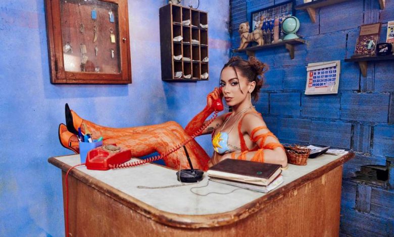 A woman with styled hair and bright makeup sits on a desk in a vividly blue room, talking on a red rotary phone. She wears an orange, patterned outfit inspired by Anitta's '
