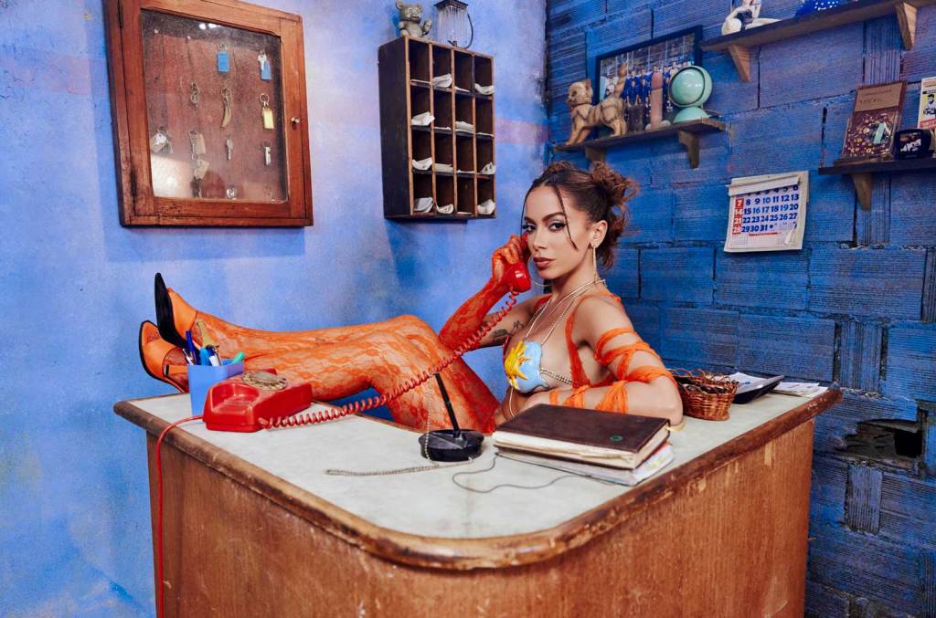 A woman with styled hair and bright makeup sits on a desk in a vividly blue room, talking on a red rotary phone. She wears an orange, patterned outfit inspired by Anitta's '