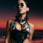 A woman with sleek sunglasses and glossy lips wears a futuristic black and blue outfit with a chunky chain necklace. she stands before a vibrant sunset sky and blurry city lights in the background.
