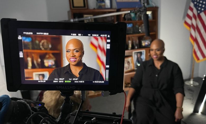 A video camera's LCD screen displays a live recording of a bald black woman smiling in a black shirt, seated in a studio with a blurred American flag and bookshelf in the background. The screen shows