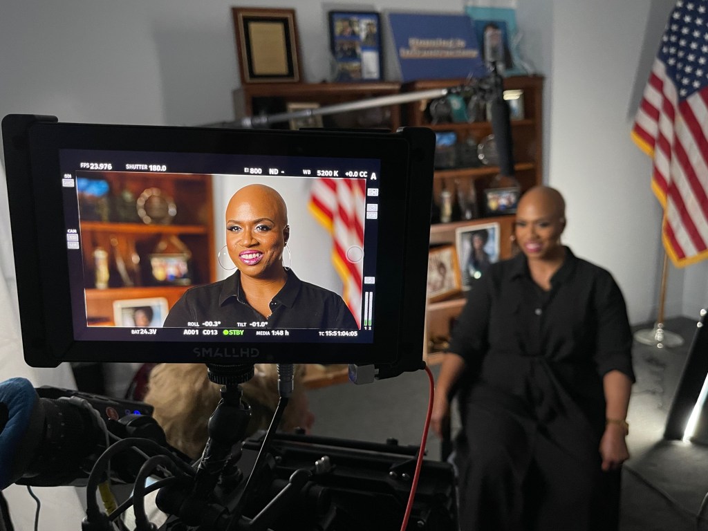 A video camera's LCD screen displays a live recording of a bald black woman smiling in a black shirt, seated in a studio with a blurred American flag and bookshelf in the background. The screen shows