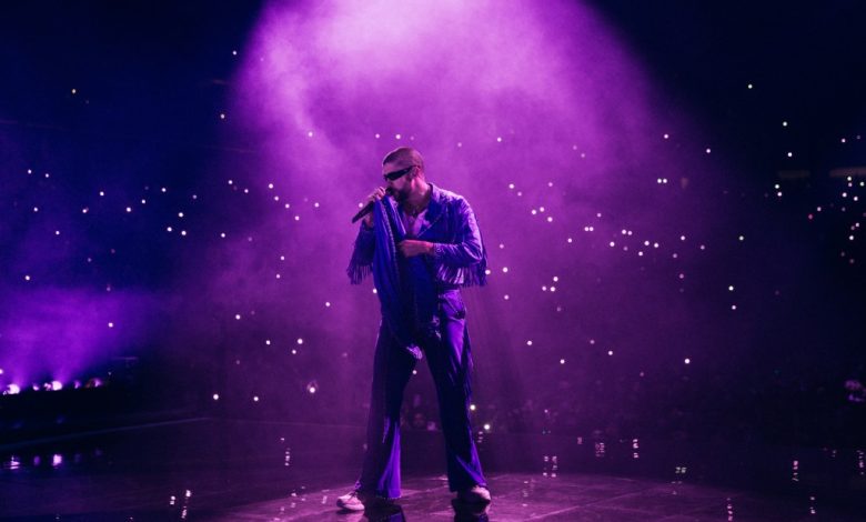 A male singer performs on stage under purple lighting, surrounded by a hazy atmosphere and cell phone lights from the audience, wearing a shiny blue jacket and sunglasses at Bad Bunny's Most Wanted Tour.