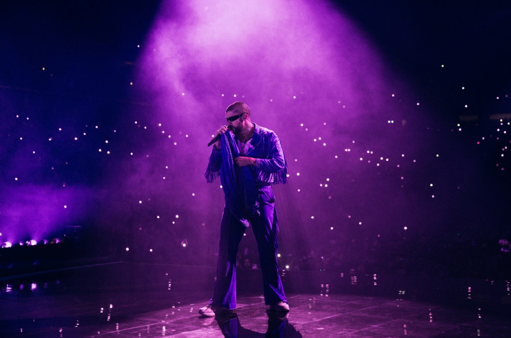 A male singer performs on stage under purple lighting, surrounded by a hazy atmosphere and cell phone lights from the audience, wearing a shiny blue jacket and sunglasses at Bad Bunny's Most Wanted Tour.
