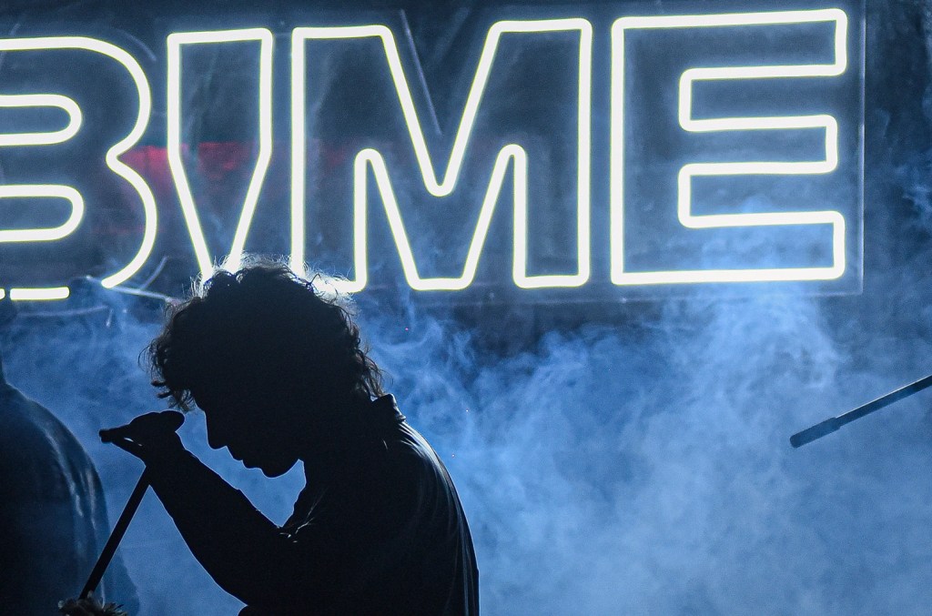 Silhouetted figure of a person singing into a microphone on a foggy stage, with a bright neon sign that reads "BIME Bogotá 2024" in the background, creating