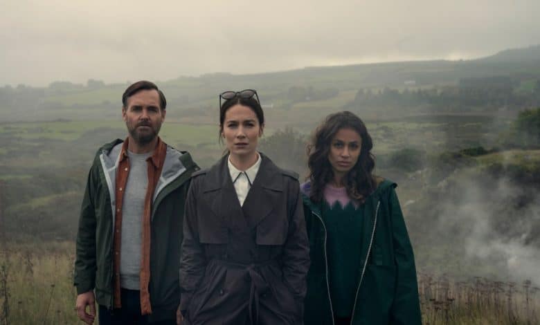 Three people stand in a misty rural landscape. A man on the left wears a green jacket, a woman in the center has on a gray trench coat and glasses, and another woman on the right