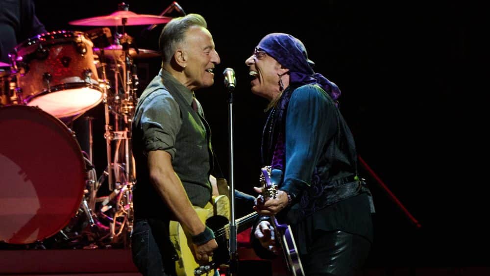 Two musicians on stage engaging enthusiastically; one plays the guitar and the other sings into a microphone, both smiling widely near a drum set under a spotlight during Springsteen's epic show.