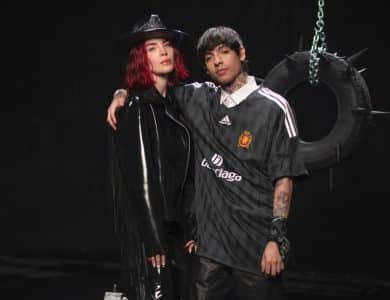 Two people, one with red hair in a black hat and shiny trench coat, and another with dark hair in a manchester united jersey, stand together in front of a tire swing hanging on a chain, against a dark background.