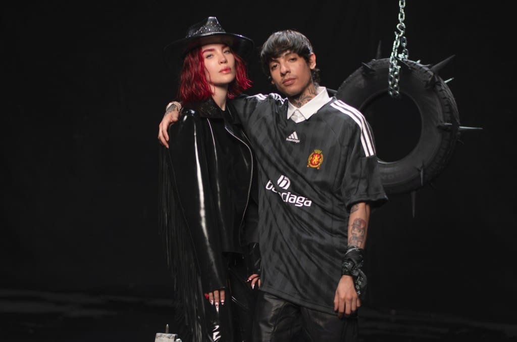 Two people, one with red hair in a black hat and shiny trench coat, and another with dark hair in a manchester united jersey, stand together in front of a tire swing hanging on a chain, against a dark background.