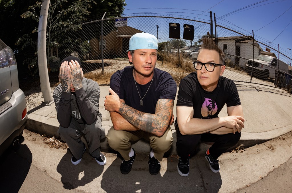 Three men posing outdoors, one covering his face with his hands, the middle one with tattooed arms crossed, and the third wearing glasses. They are wearing casual attire, with urban scenery and a Blink