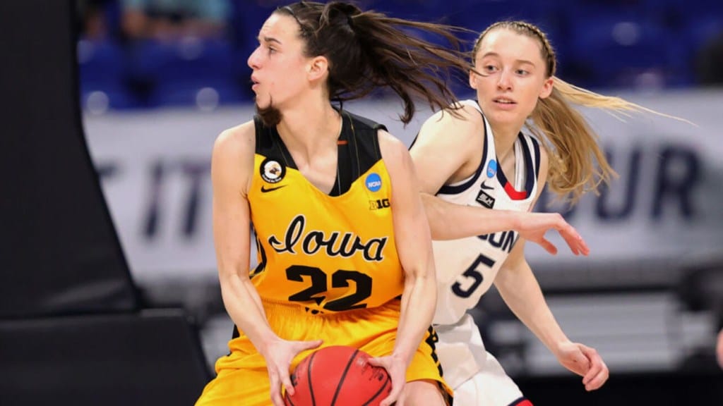 Two female basketball players in motion during a game. The player in a yellow Iowa jersey, number 22, dribbles the ball, while a player in a white UConn jersey from a rival team