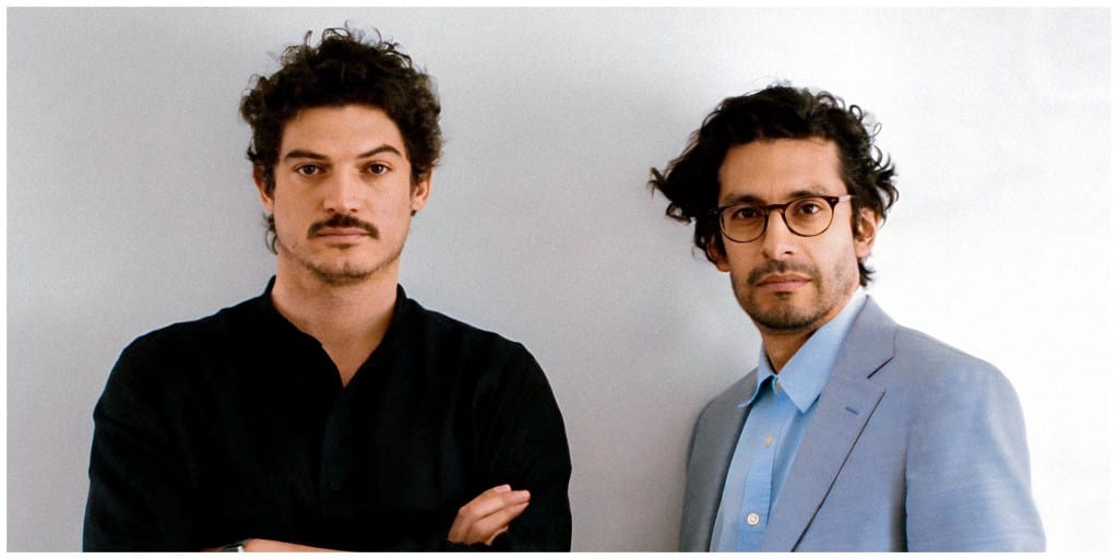 Two men standing against a plain background; one in a black shirt with crossed arms, the other in a light blue blazer looking serious. Both are Colombian filmmakers with curly hair and wear glasses.