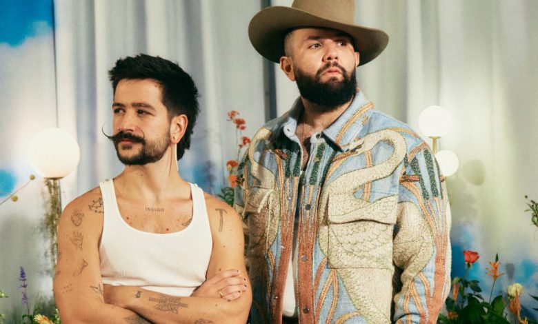 Two men pose confidently, one in a white tank top with tattoos, and the other in a patterned jacket and cowboy hat, channeling the Aventura vibe, in a room with soft lighting