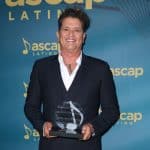 Carlos Vives, in a black suit and open-collar white shirt, smiles while holding a glass award at the ASCAP Latin Awards. The background is blue with the ASCAP logo and musical notes pattern