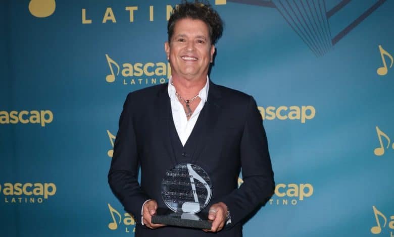 Carlos Vives, in a black suit and open-collar white shirt, smiles while holding a glass award at the ASCAP Latin Awards. The background is blue with the ASCAP logo and musical notes pattern