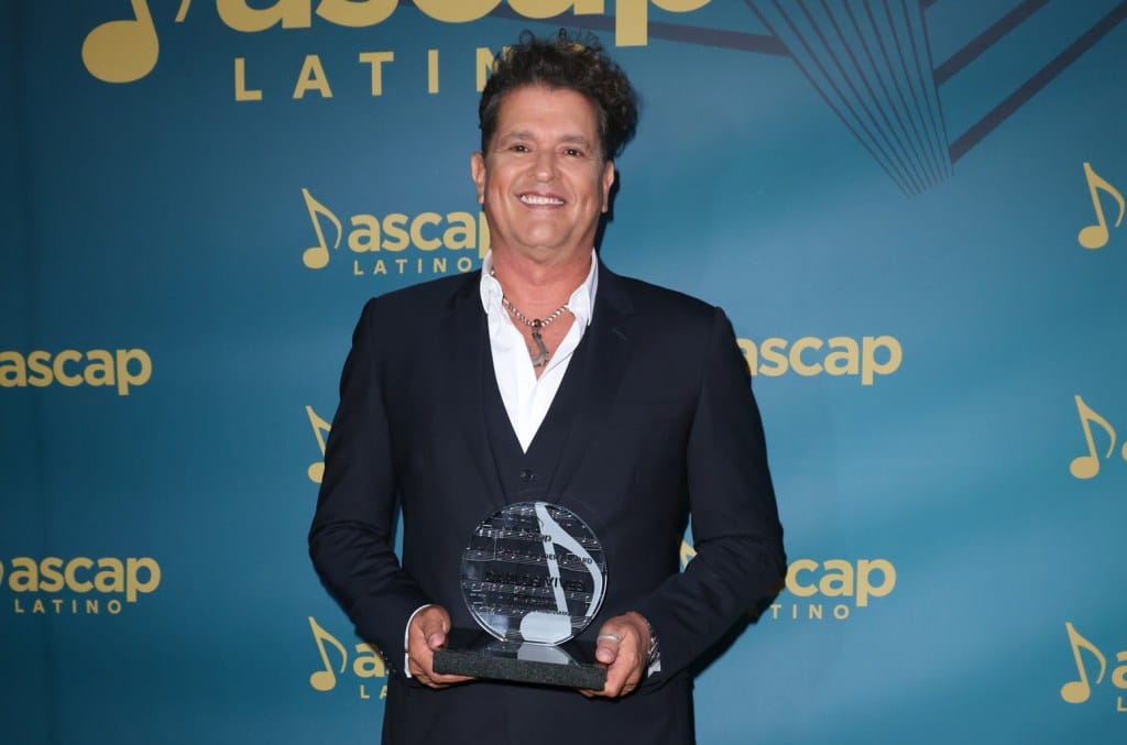 Carlos Vives, in a black suit and open-collar white shirt, smiles while holding a glass award at the ASCAP Latin Awards. The background is blue with the ASCAP logo and musical notes pattern