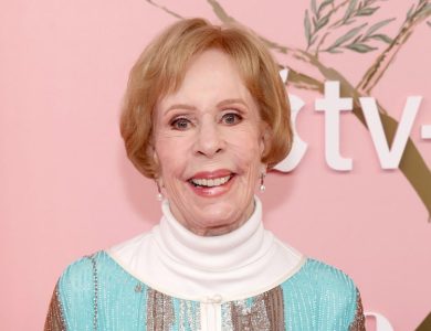 Carol Burnett smiles at an event, wearing a turquoise sequined dress with a high white collar. She stands against a pink backdrop with decorative elements, and a logo for "tv+" is visible.
