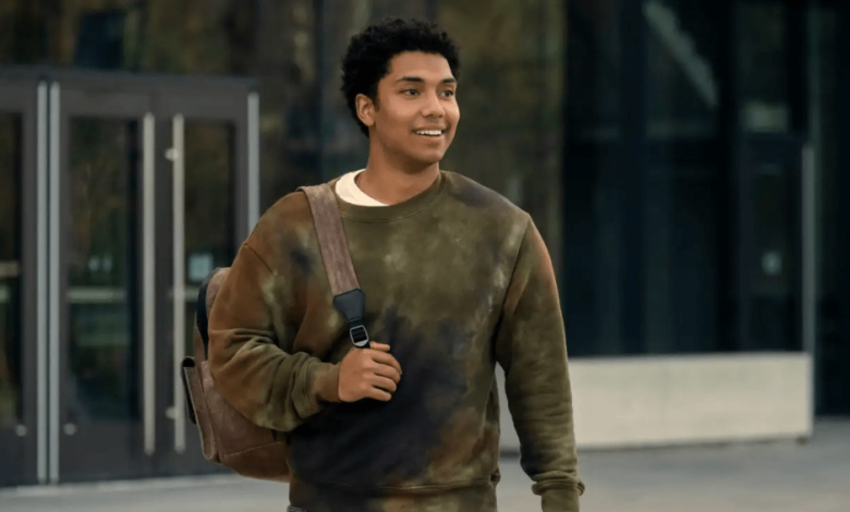 A young man with curly hair and a light beard smiles while walking. He wears a green sweater and carries a shoulder bag, with a modern building in the background.