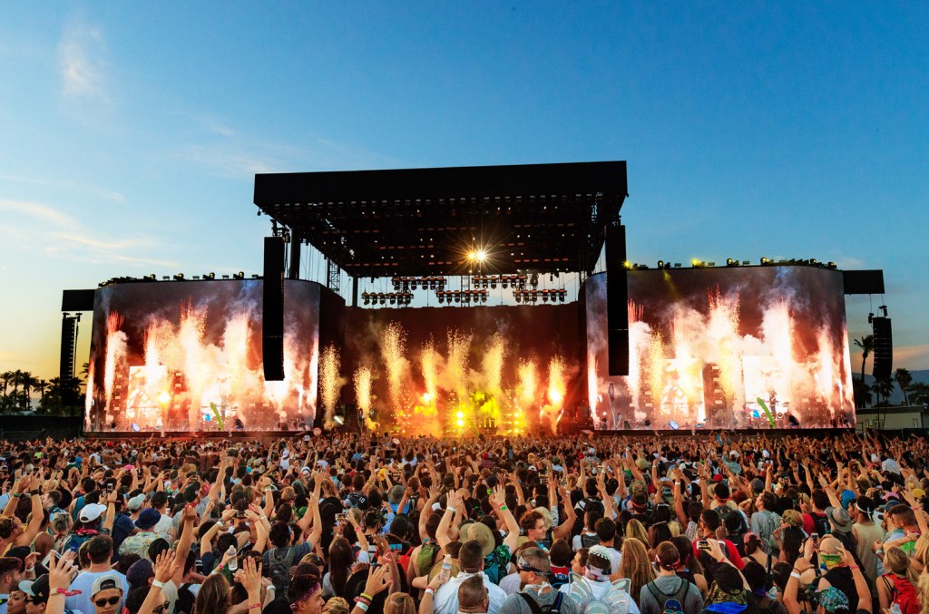 A large crowd at a music festival enjoying a live performance. The stage features dynamic lighting, smoke effects, and large screens displaying the band. The sky above is at dusk, adding a dramatic
