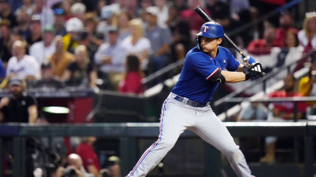 A baseball player in a blue and red Texas Rangers uniform prepares to swing at a pitch during a 2024 MLB game, with spectators blurred in the background.