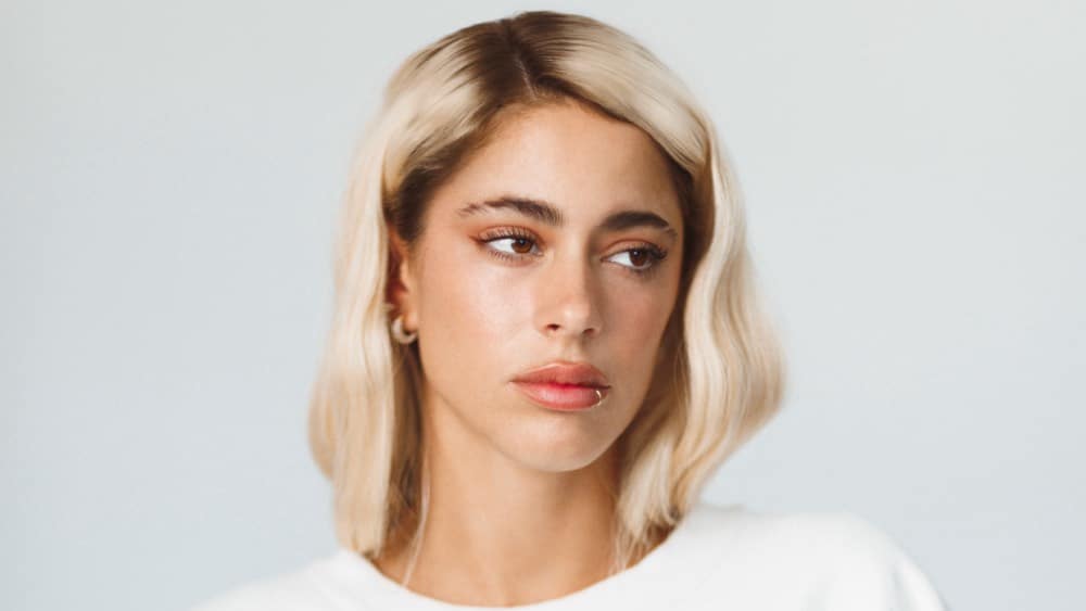 A young woman with shoulder-length blond hair and dark roots, featuring soft waves, embodies the cover of Tini's intimate album 'Un Mechón de Pelo'. She has light skin, prominent eyebrows