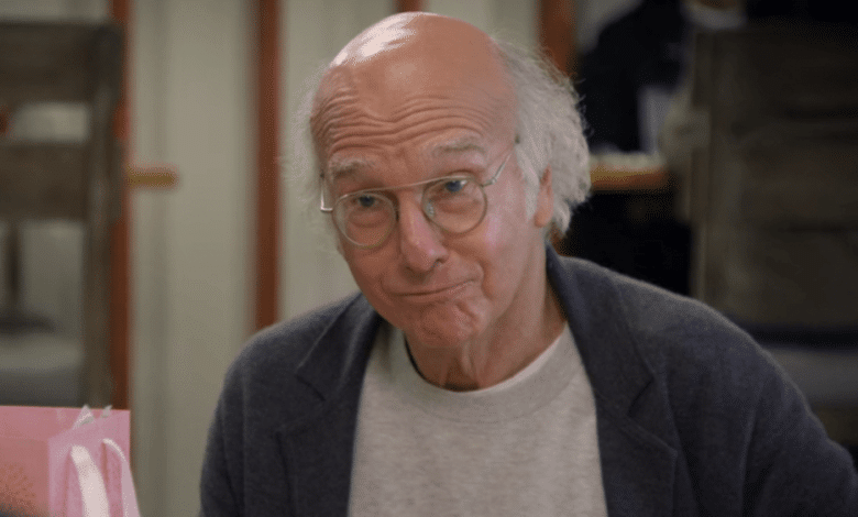An older bald man with slight stubble and glasses looks directly at the camera, making a humorous face reminiscent of Larry David. He's raising his eyebrows and has a subtle smile, wearing a grey sweater
