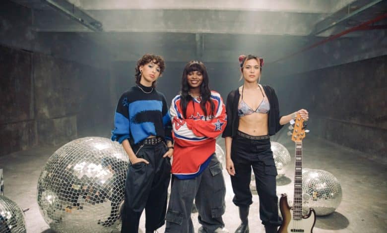 Three young women stand confidently in a concrete room with large disco balls. The person on the left wears a blue-and-black sweater, the middle person sports a red jersey, and the one on the right