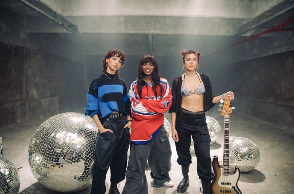 Three young women stand confidently in a concrete room with large disco balls. The person on the left wears a blue-and-black sweater, the middle person sports a red jersey, and the one on the right