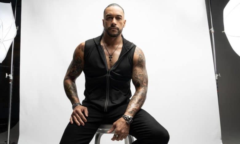 Damian Priest, with tattoos on his arms, sits on a stool between two photography umbrellas, wearing a black sleeveless shirt and vest, looking confidently at the camera.