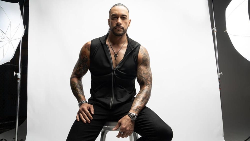 Damian Priest, with tattoos on his arms, sits on a stool between two photography umbrellas, wearing a black sleeveless shirt and vest, looking confidently at the camera.